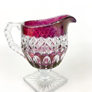 May include: A clear glass creamer with a ruby red top and a cut glass design. The creamer has a square base and a handle.