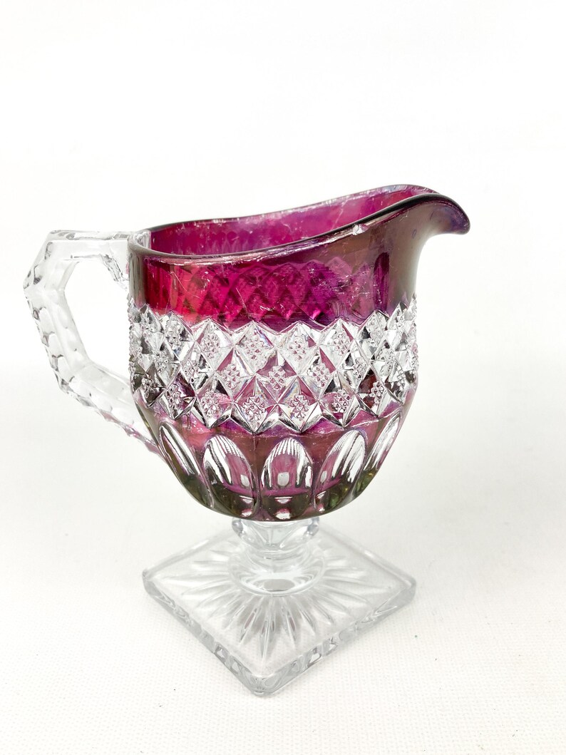May include: A clear glass creamer with a ruby red top and a cut glass design. The creamer has a square base and a handle.