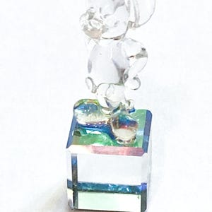 May include: A clear glass figurine of a cartoon mouse standing on a square base. The mouse is posed with one hand raised. The base has a colorful, iridescent surface.
