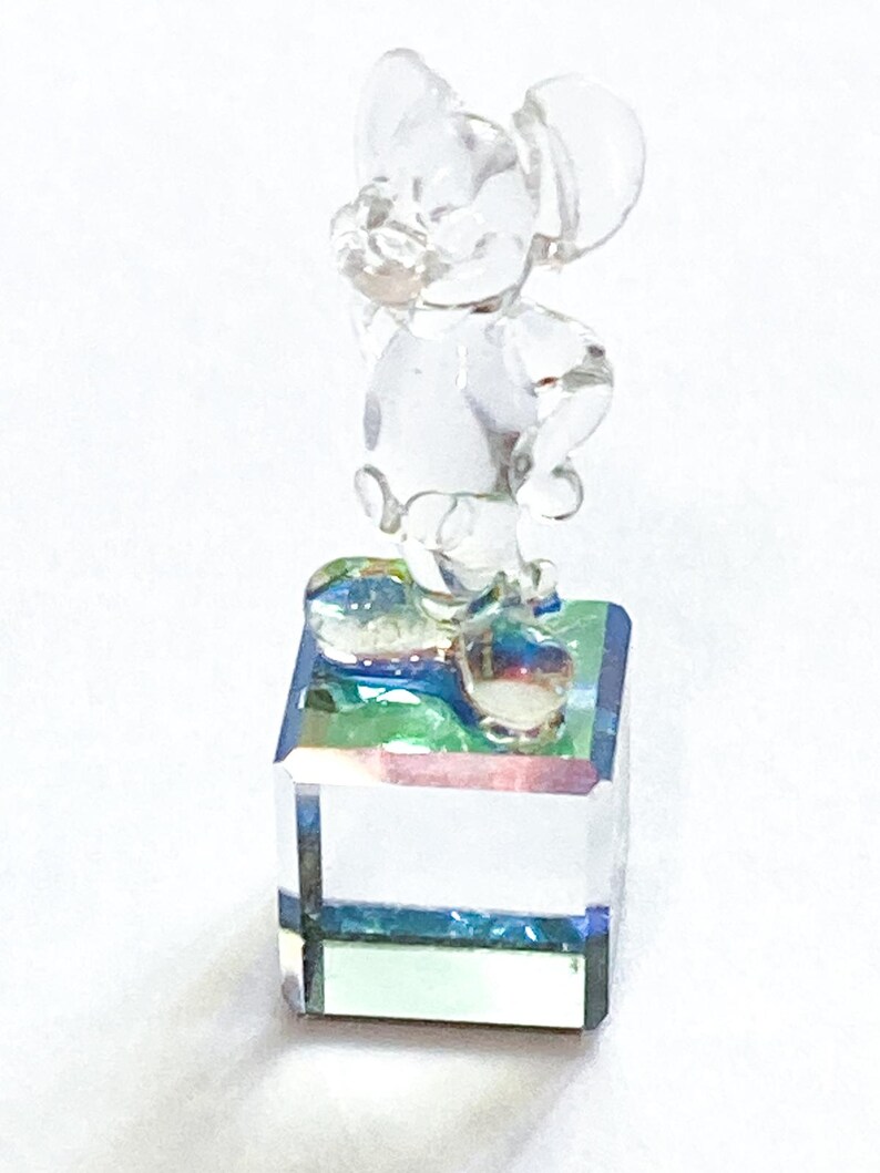 May include: A clear glass figurine of a cartoon mouse standing on a square base. The mouse is posed with one hand raised. The base has a colorful, iridescent surface.