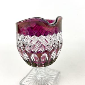 May include: A clear glass pitcher with a ruby red base and a clear, diamond-shaped pattern. The pitcher has a square base and a wide, rounded top.