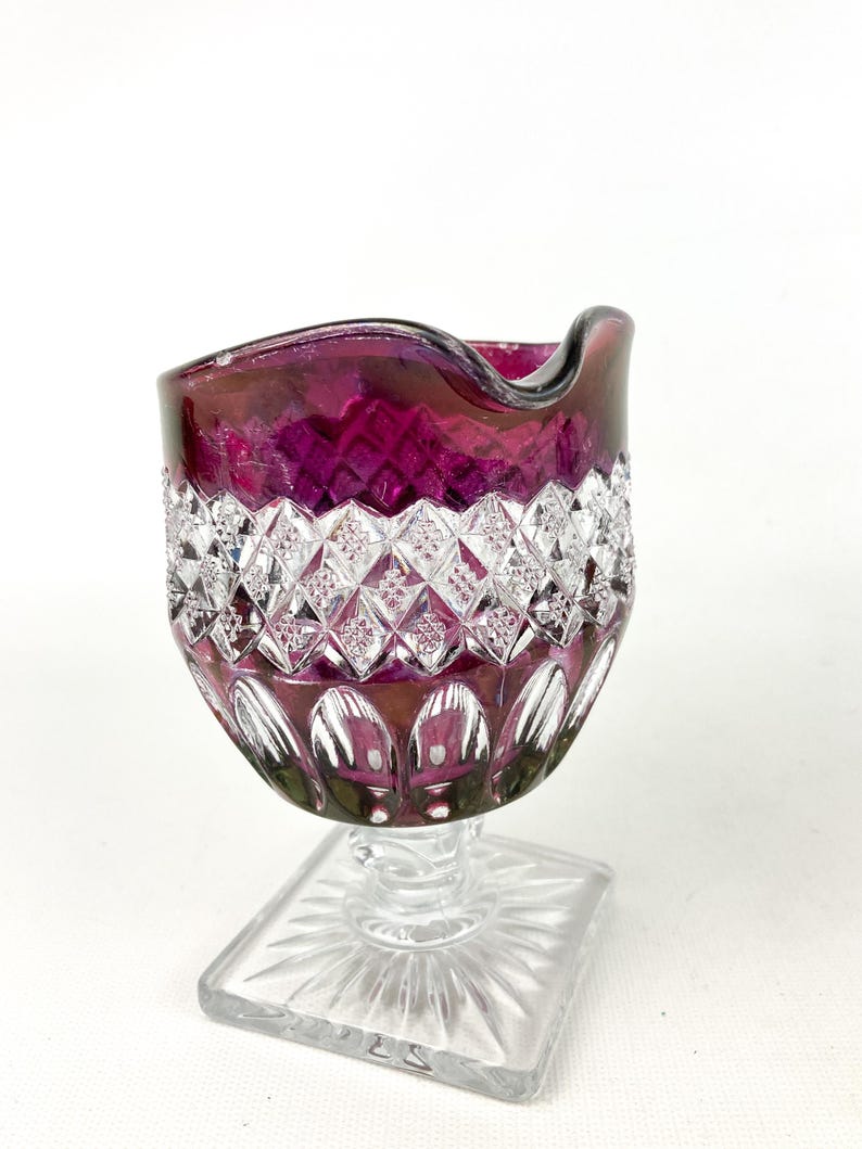 May include: A clear glass pitcher with a ruby red base and a clear, diamond-shaped pattern. The pitcher has a square base and a wide, rounded top.