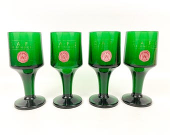 May include: Four green glass shot glasses with gold lettering that reads "Cafe De Benedictine" and a red crest. The glasses have a wide base and a narrow stem.