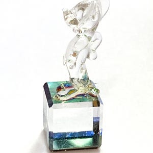 May include: A clear glass figurine of a cat standing on a square, faceted base. The base has a blue and green iridescent effect. The cat is posed with its head tilted and ears perked up, with a playful appearance.