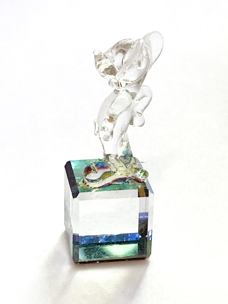 May include: A clear glass figurine of a cat standing on a square, faceted base. The base has a blue and green iridescent effect. The cat is posed with its head tilted and ears perked up, with a playful appearance.