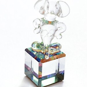 May include: A clear glass mouse figurine with large ears and arms akimbo, perched atop a square, iridescent glass block. The figurine is transparent, allowing light to pass through, creating a shimmering effect. A decorative item.