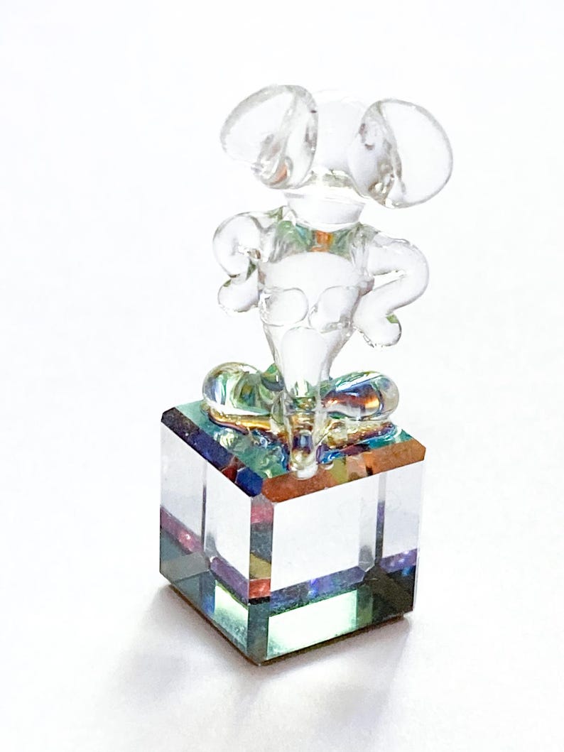 May include: A clear glass mouse figurine with large ears and arms akimbo, perched atop a square, iridescent glass block. The figurine is transparent, allowing light to pass through, creating a shimmering effect. A decorative item.