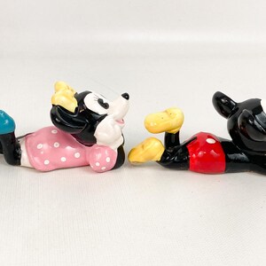 May include: Two ceramic figurines of Mickey and Minnie Mouse in a relaxed pose. Minnie wears a pink polka-dot dress and blue shoes. Mickey has red shorts and yellow shoes. Both have black and white features.