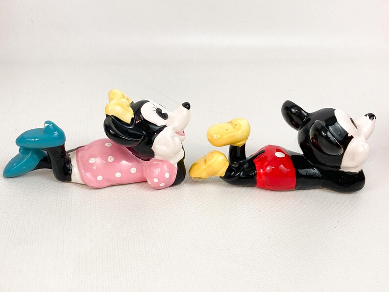 May include: Two ceramic figurines of Mickey and Minnie Mouse in a relaxed pose. Minnie wears a pink polka-dot dress and blue shoes. Mickey has red shorts and yellow shoes. Both have black and white features.