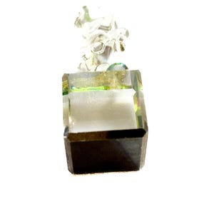 May include: A clear, faceted crystal pendant with a square base and a detailed, abstract design at the top. The crystal has a dark brown bottom and a light green and gold interior, creating a unique visual effect. The pendant is suspended from a silver-colored bail.