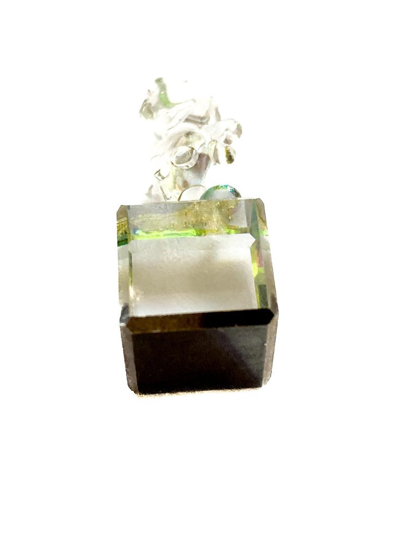 May include: A clear, faceted crystal pendant with a square base and a detailed, abstract design at the top. The crystal has a dark brown bottom and a light green and gold interior, creating a unique visual effect. The pendant is suspended from a silver-colored bail.