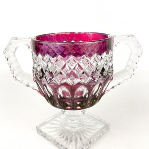 May include: A clear glass sugar bowl with two handles and a square base. The bowl is decorated with a diamond pattern and has a deep purple hue.