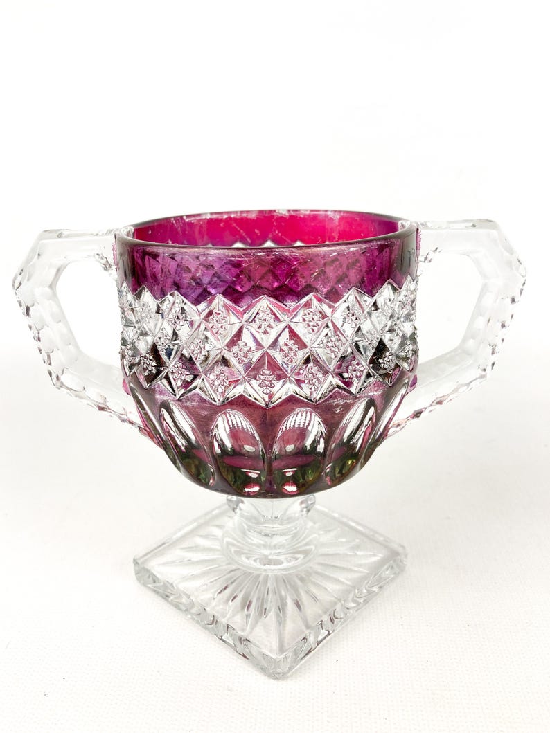 May include: A clear glass sugar bowl with two handles and a square base. The bowl is decorated with a diamond pattern and has a deep purple hue.