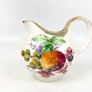 May include: A white ceramic creamer with a gold rim and a fruit design. The design features a red and orange apple, green grapes, and blackberries.
