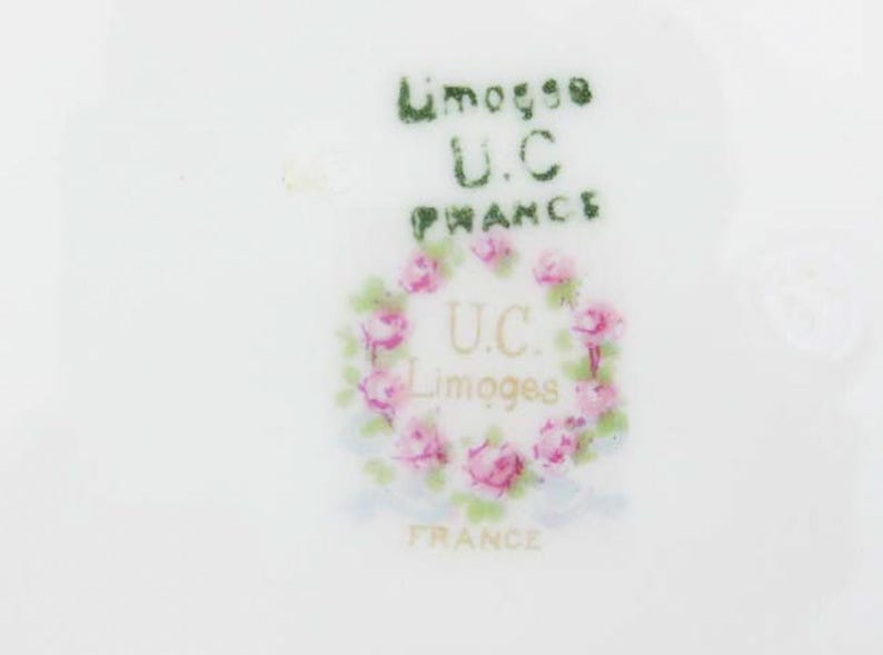 May include: Close-up of a white porcelain surface with the text "Limoges U.C. France" in green and gold. A wreath of pink roses encircles the letters "U.C. Limoges France".