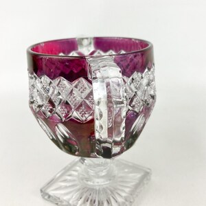 May include: A clear glass goblet with a square base and a deep purple rim. The goblet has a cut glass design and a clear glass handle.