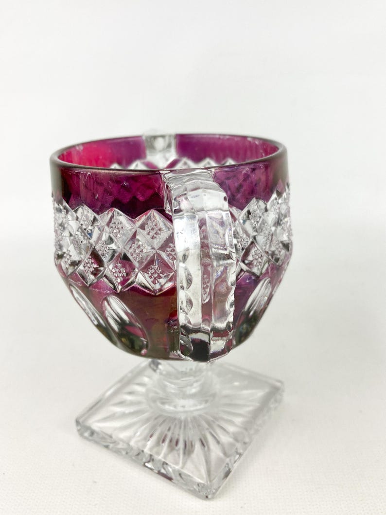 May include: A clear glass goblet with a square base and a deep purple rim. The goblet has a cut glass design and a clear glass handle.
