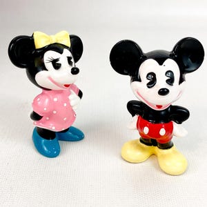 Może przedstawiać: Dwie figurki postaci z kresk&oacute;wek Myszki Miki i Myszki Minnie. Myszka Minnie ma na sobie r&oacute;żową sukienkę w kropki, niebieskie buty i ż&oacute;łtą kokardę. Myszka Miki ma na sobie czerwone szorty, ż&oacute;łte buty i czarny top.