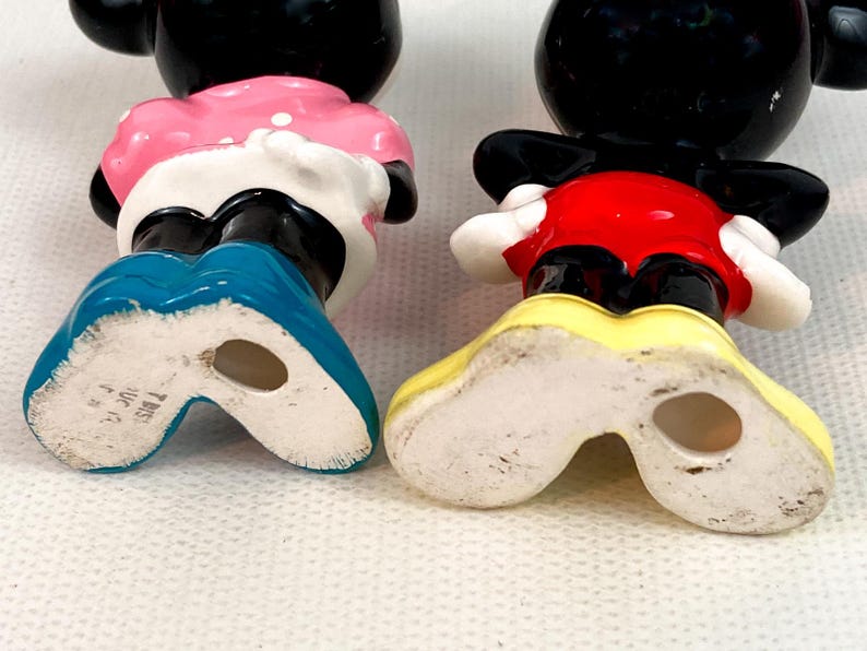 Może przedstawiać: Para zabytkowych figurek Myszki Miki i Myszki Minnie. Miki ma na sobie czerwone szorty i ż&oacute;łte buty. Myszka Minnie ma na sobie r&oacute;żową sukienkę w białe kropki i niebieskie buty. Obie mają czarne głowy i białe rękawiczki.