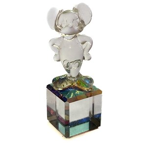 May include: A clear glass figurine of a mouse with large ears, standing with hands on hips. The mouse is mounted on a square, multi-colored glass base. The figurine is a decorative item.