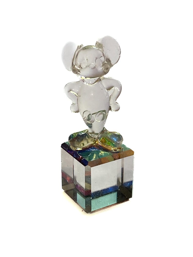 May include: A clear glass figurine of a mouse with large ears, standing with hands on hips. The mouse is mounted on a square, multi-colored glass base. The figurine is a decorative item.