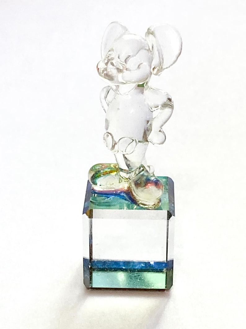 May include: A clear glass figurine of a mouse with large ears, standing on a square, iridescent base. The mouse is in a relaxed pose, with its hands on its hips. The base has blue and green hues.