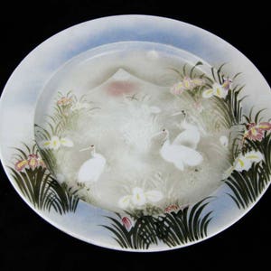 May include: A white ceramic plate with a blue rim and a painted design of white cranes, flowers, and a mountain. The plate has a light blue background with a white center.