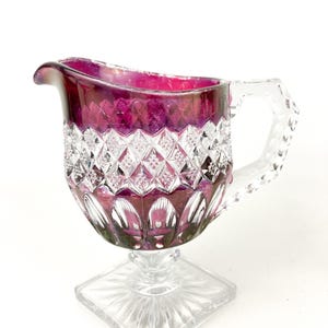 May include: A clear glass creamer with a cut glass design. The creamer has a purple iridescent finish and a square base.