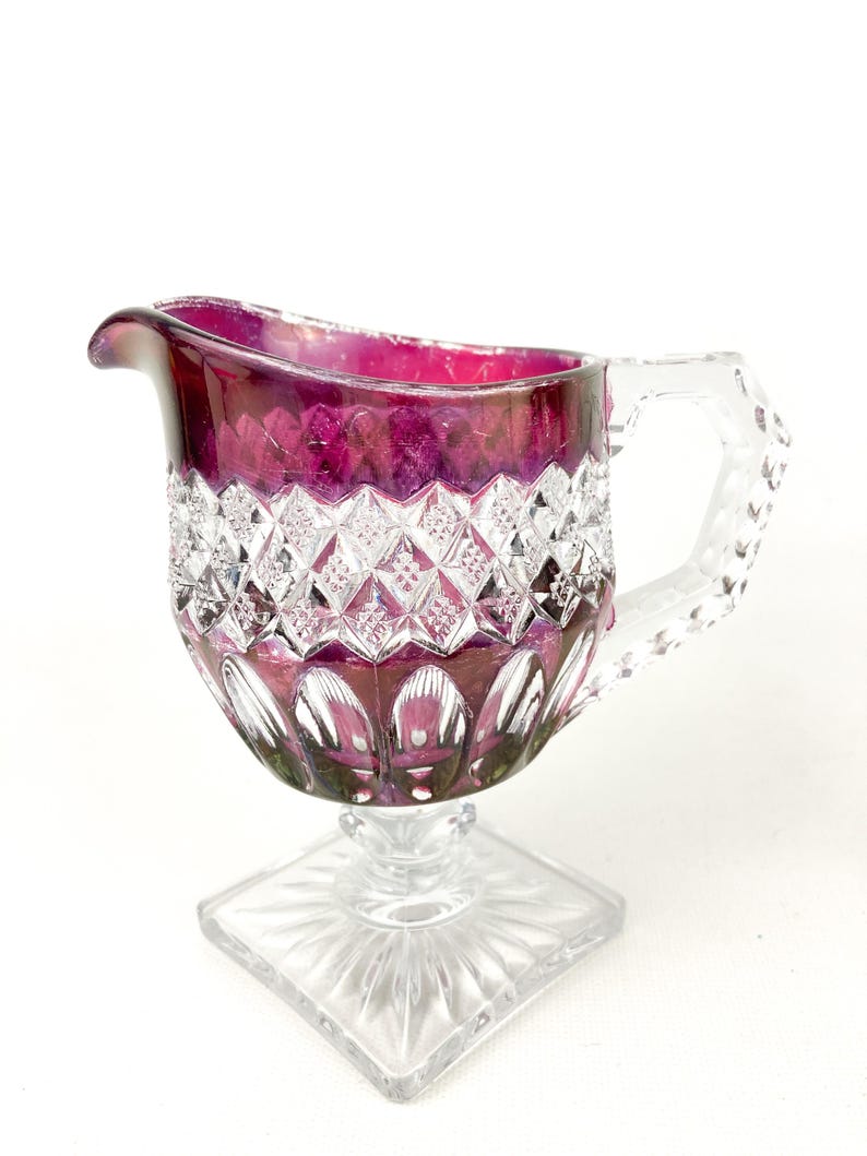 May include: A clear glass creamer with a cut glass design. The creamer has a purple iridescent finish and a square base.