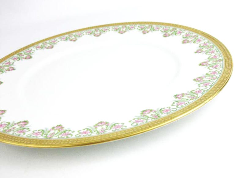 May include: Oval white serving platter with a gold rim and a floral design in pink and green. The decorative border features a repeating pattern of flowers and leaves. A classic piece for serving food.