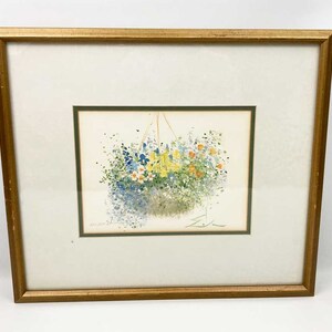 May include: A framed watercolor painting of a hanging basket overflowing with colorful flowers. The flowers are in shades of blue, yellow, and white, and the basket is made of woven material. The painting is set against a white background.