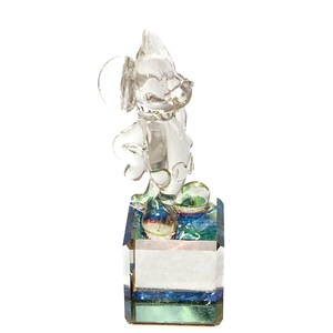 May include: A clear glass figurine of a cartoon mouse with large ears, standing on a rectangular base. The base has a layered design with blue, green, and white accents. The figurine is transparent, allowing light to pass through.