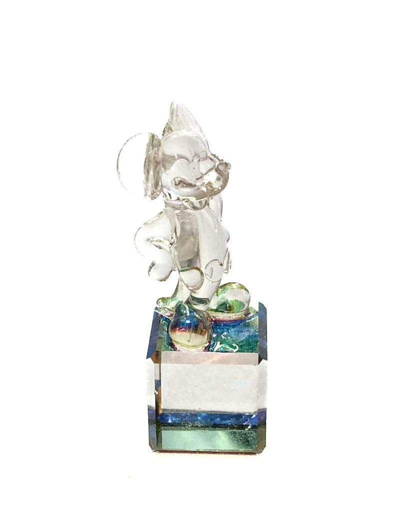 May include: A clear glass figurine of a cartoon mouse with large ears, standing on a rectangular base. The base has a layered design with blue, green, and white accents. The figurine is transparent, allowing light to pass through.