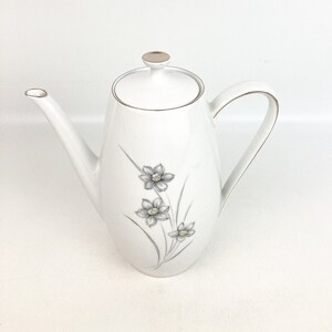 May include: White ceramic coffee pot with a floral design. The pot has a silver lid and a gold trim around the rim.