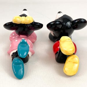 May include: Two ceramic figurines of cartoon characters, one in a pink dress with white polka dots and a yellow bow, the other in red shorts. Both have black ears and shoes. The figurines are positioned facing away from the viewer.