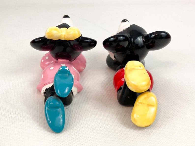 May include: Two ceramic figurines of cartoon characters, one in a pink dress with white polka dots and a yellow bow, the other in red shorts. Both have black ears and shoes. The figurines are positioned facing away from the viewer.