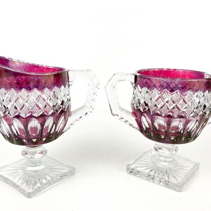 May include: A set of two vintage glass creamer and sugar bowl. The glass is clear with a ruby red iridescent finish. The creamer has a handle and a square base. The sugar bowl has two handles and a square base.