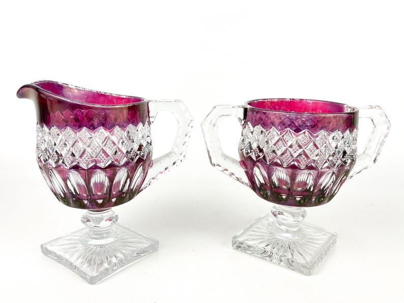 May include: A set of two vintage glass creamer and sugar bowl. The glass is clear with a ruby red iridescent finish. The creamer has a handle and a square base. The sugar bowl has two handles and a square base.