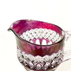 May include: A clear glass creamer with a cut glass design. The creamer is a deep purple color and has a handle.