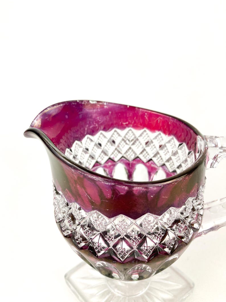 May include: A clear glass creamer with a cut glass design. The creamer is a deep purple color and has a handle.