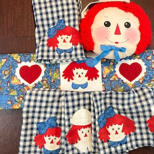 May include: A collection of handmade Raggedy Ann-themed items. Includes a round pillow with a Raggedy Ann face, blue and white checkered fabric, and appliqued Raggedy Ann faces and hearts. The items are likely kitchen or home decor.