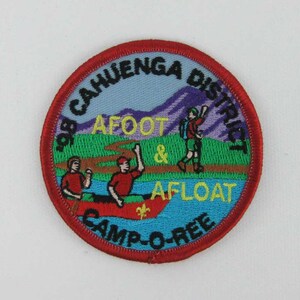 May include: A red, white, and blue embroidered patch with the text "1998 CAHUENGA DISTRICT CAMP-O-REE AFOOT & AFLOAT". The patch features a mountain range, a hiker, and two people in a canoe.