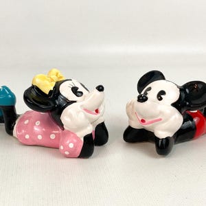 May include: Two ceramic figurines of Disney characters, Mickey and Minnie Mouse, are lying down. Minnie wears a pink polka-dot dress, a yellow bow, and blue shoes. Mickey wears red shorts and yellow shoes. Both have black ears.