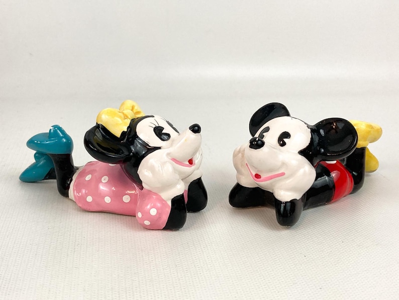 May include: Two ceramic figurines of Disney characters, Mickey and Minnie Mouse, are lying down. Minnie wears a pink polka-dot dress, a yellow bow, and blue shoes. Mickey wears red shorts and yellow shoes. Both have black ears.