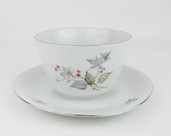 May include: A white porcelain gravy boat with a floral design featuring green leaves and pink flowers. The boat is sitting on a matching saucer with a silver rim.