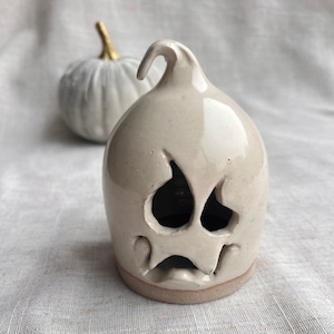 Peut inclure: Une figurine de fantôme en céramique blanc cassé brillant avec un sommet incurvé et des traits du visage découpés, incluant deux grandes orbites et une large bouche tombante. La base est non émaillée, révélant une argile beige, avec une citrouille.