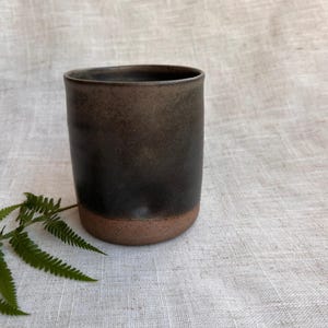 Peut inclure: Un gobelet en céramique fait à la main, avec un extérieur brun foncé texturé et une base brun rougeâtre. La tasse cylindrique est adaptée aux boissons et présente un design simple et moderne. Le fond est un tissu de lin neutre.