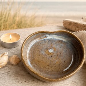 May include: A heart-shaped ceramic dish with a speckled brown and blue glaze. The bowl features a smooth, glossy finish with a subtle white center accent. The edges are rounded and slightly raised, creating a shallow basin for small items.
