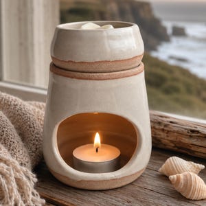 May include: A ceramic wax warmer with a cream-colored glaze and exposed terracotta-toned clay edges. The structure features a top bowl for wax melts and a lower arched opening containing a lit tea light candle, creating a warm, soft glow.