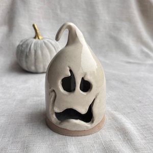 May include: A glossy, off-white ceramic ghost figurine with a curved top and cut-out facial features including two oval eyes and a wide, open mouth. Behind the ghost sits a small, pale grey pumpkin with a gold-painted stem.
